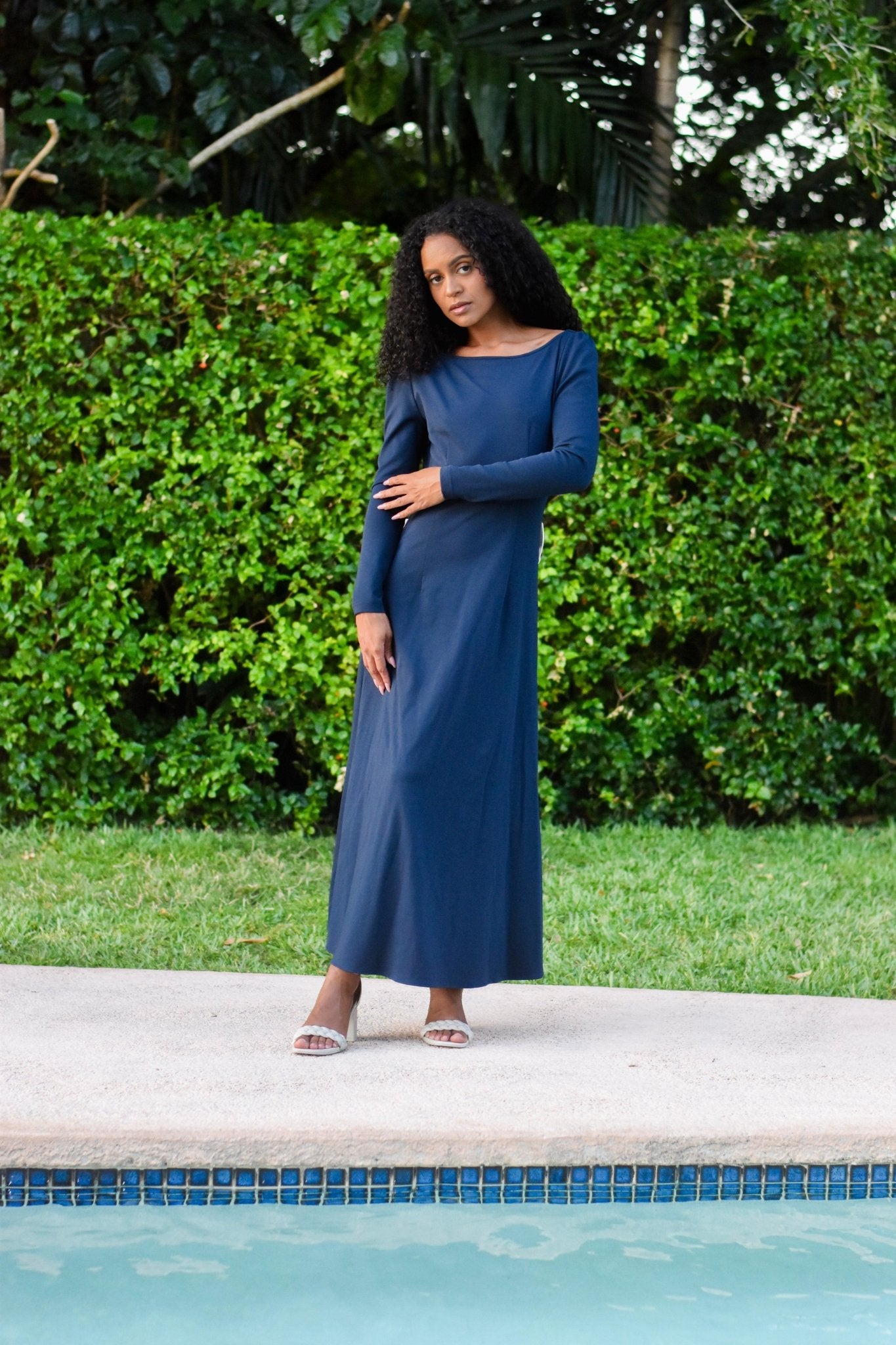 Elegant model wearing the navy blue Pansy gown by the pool, showcasing its luxurious drape and open back design.