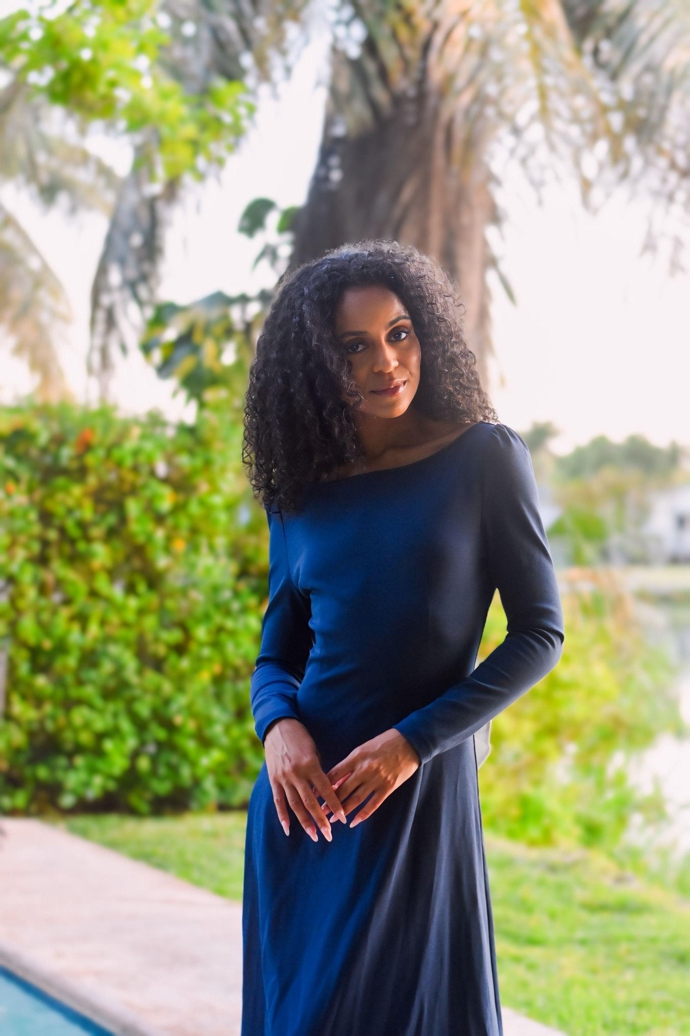 Elegant woman wearing the royal navy Pansy gown by a serene outdoor setting with palm trees.