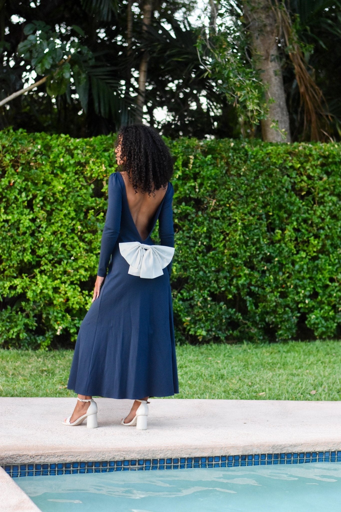 Model wearing the Pansy gown in royal navy blue with an open back and detachable bow, by the poolside.