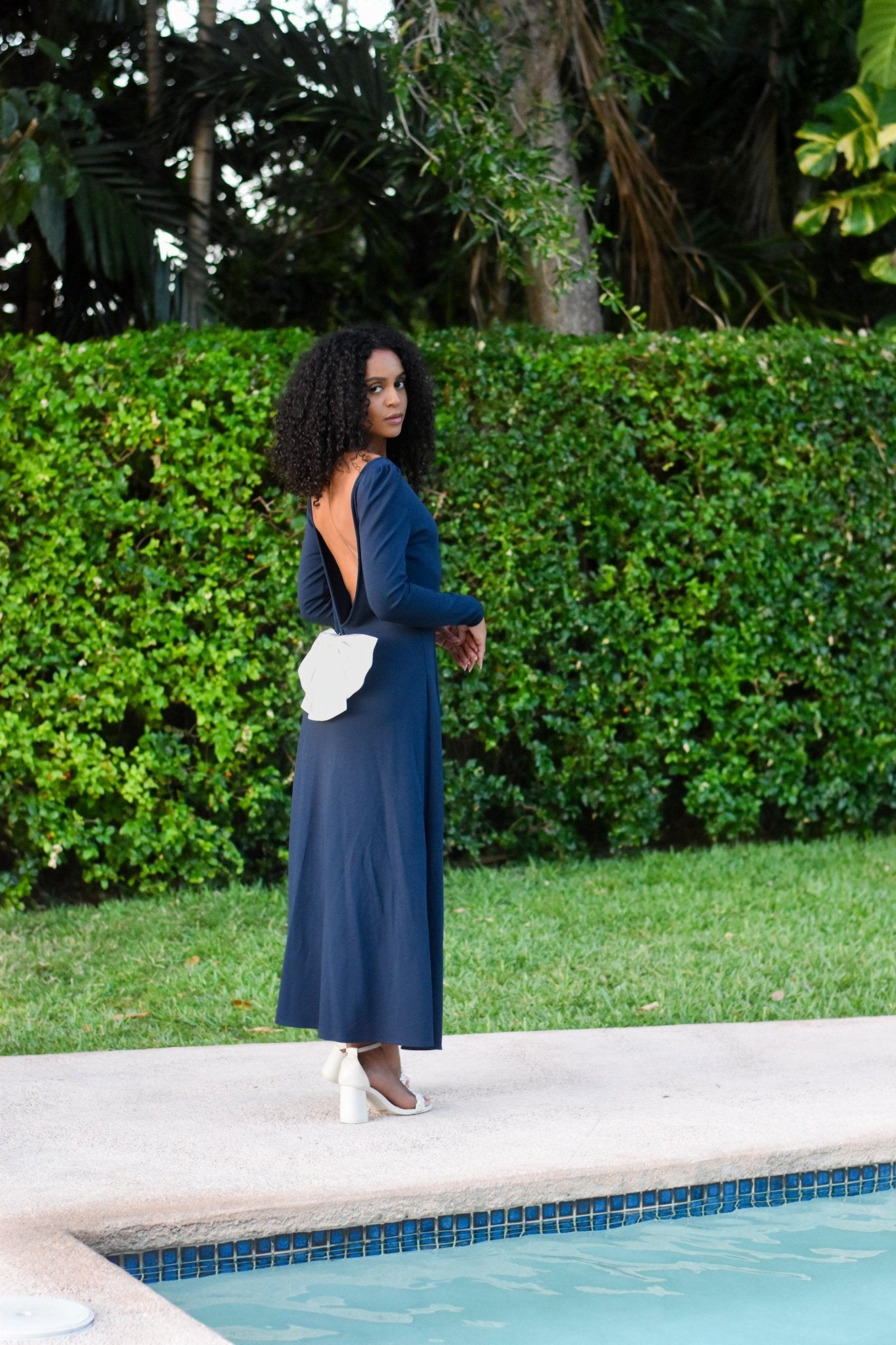 Model wearing the elegant navy blue Pansy gown with an open back, featuring a detachable bow, by a poolside.