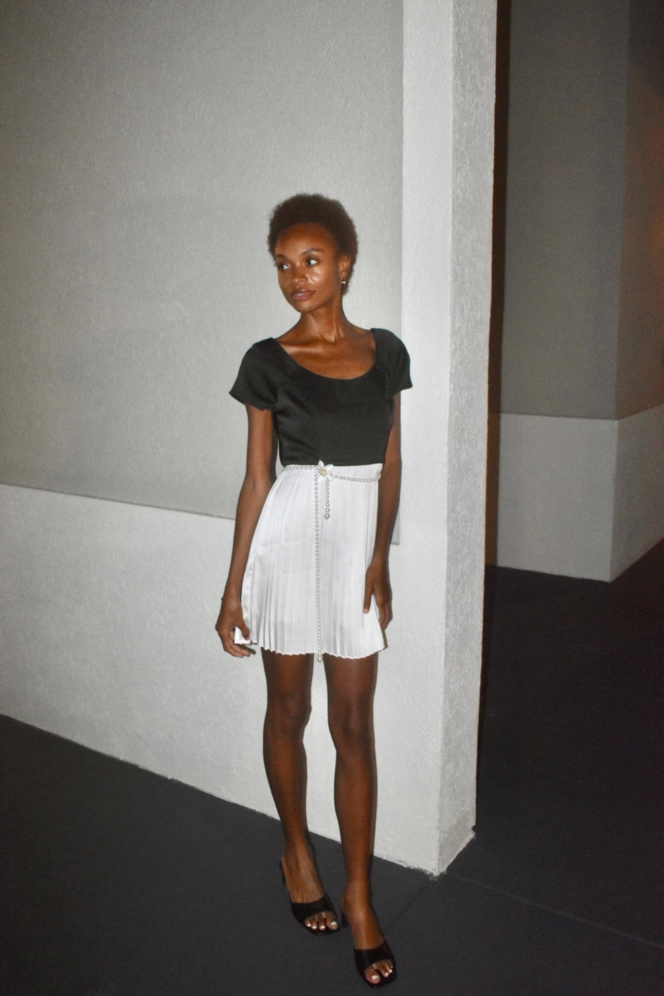 Model wearing The Viviene Dress, featuring a black top and white pleated satin skirt against a modern backdrop.